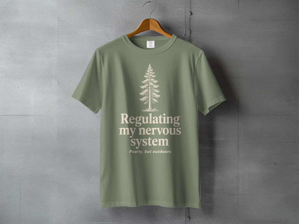 Sage green t-shirt with a minimalist pine tree and "Regulating my nervous system poorly, but outdoors" text.