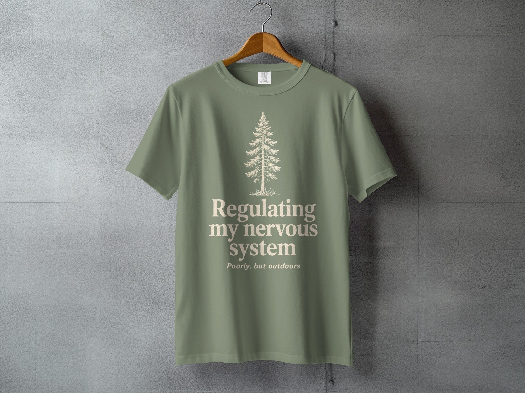 Sage green t-shirt with a minimalist pine tree and "Regulating my nervous system poorly, but outdoors" text.