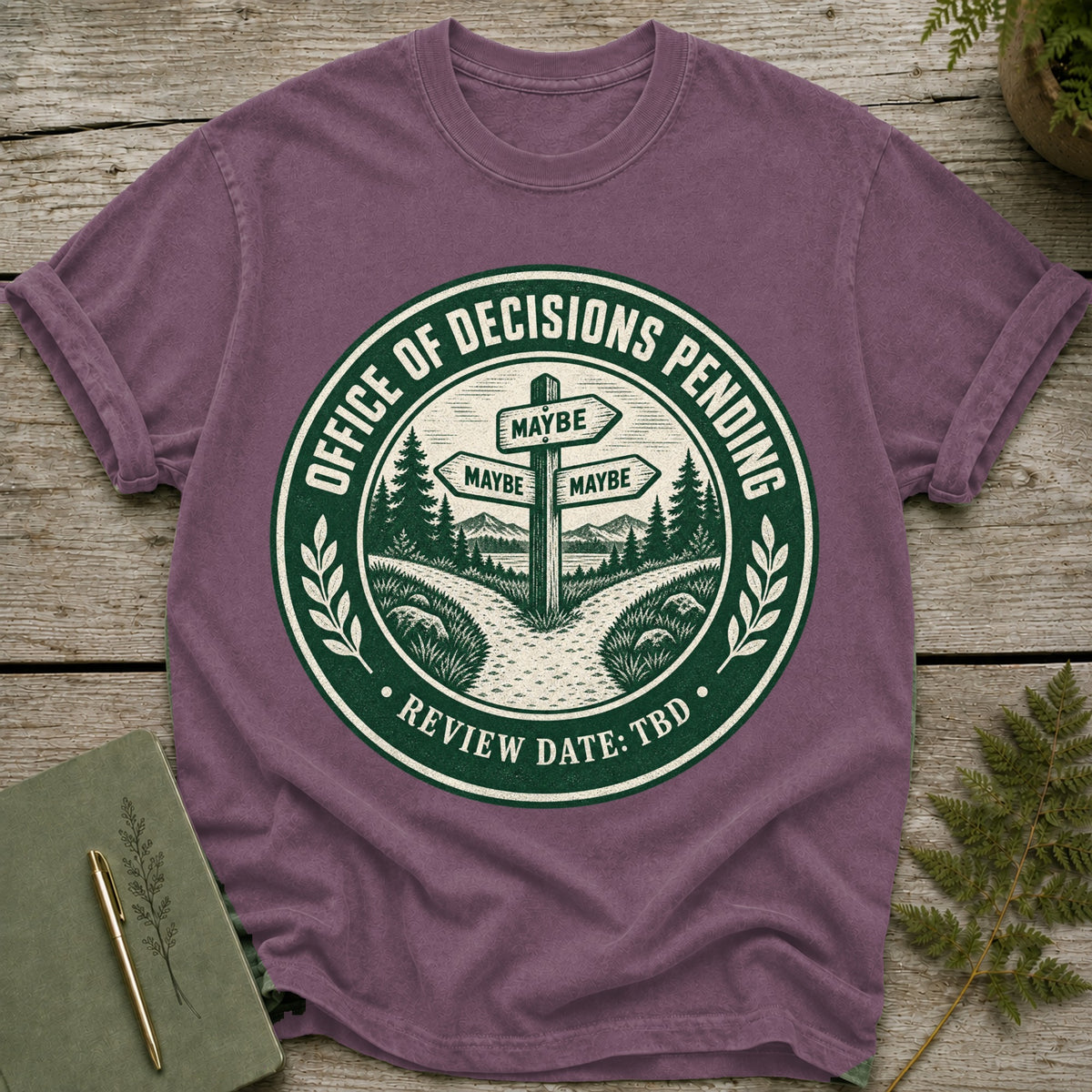 Purple t-shirt with a green circular graphic depicting a signpost with "MAYBE" signs, trees, and mountains.