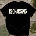 Black t-shirt with "RECHARGING" text in cream, on a wooden surface.