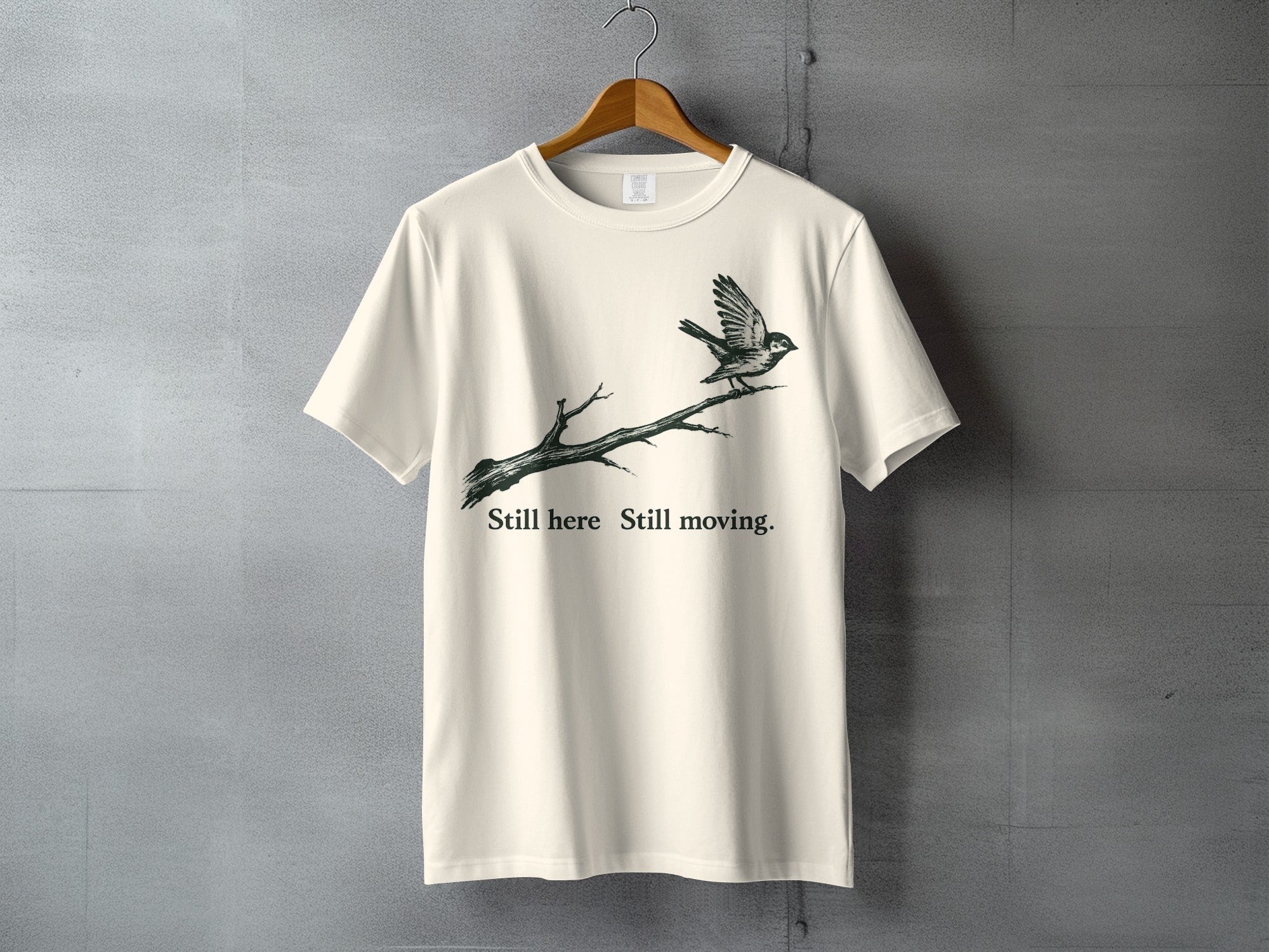 Cream t-shirt with graphic of bird on a branch and text "Still here Still moving."