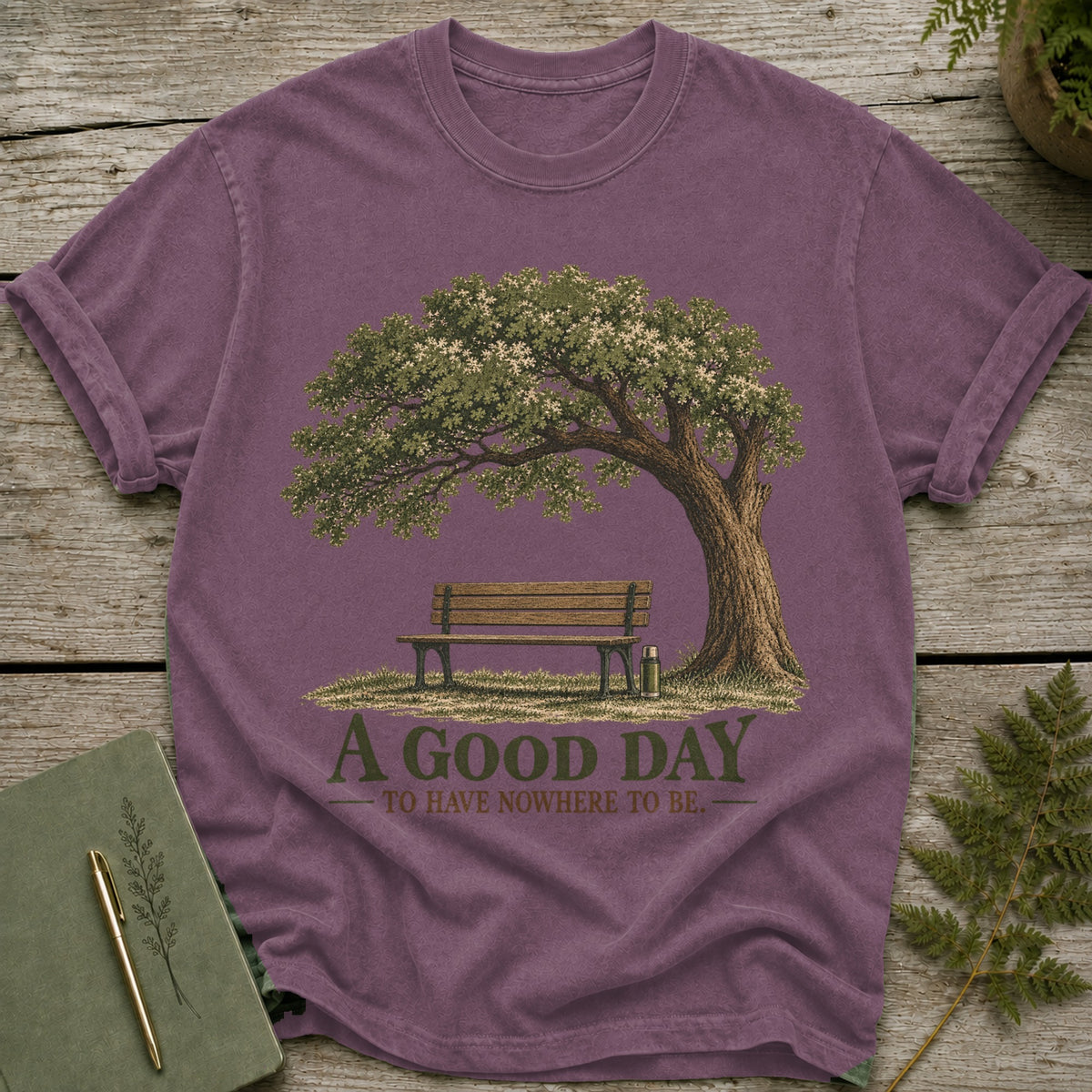 Purple t-shirt with illustration of a tree, bench, and thermos, featuring text "A GOOD DAY TO HAVE NOWHERE TO BE."