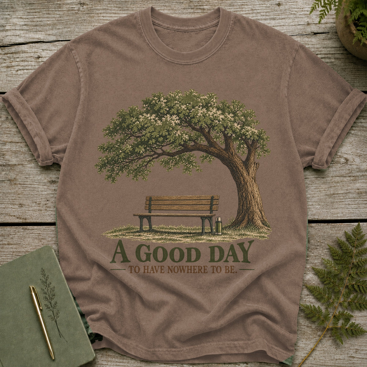 Brown t-shirt with illustration of a tree, bench, and thermos with text "A good day to have nowhere to be."
