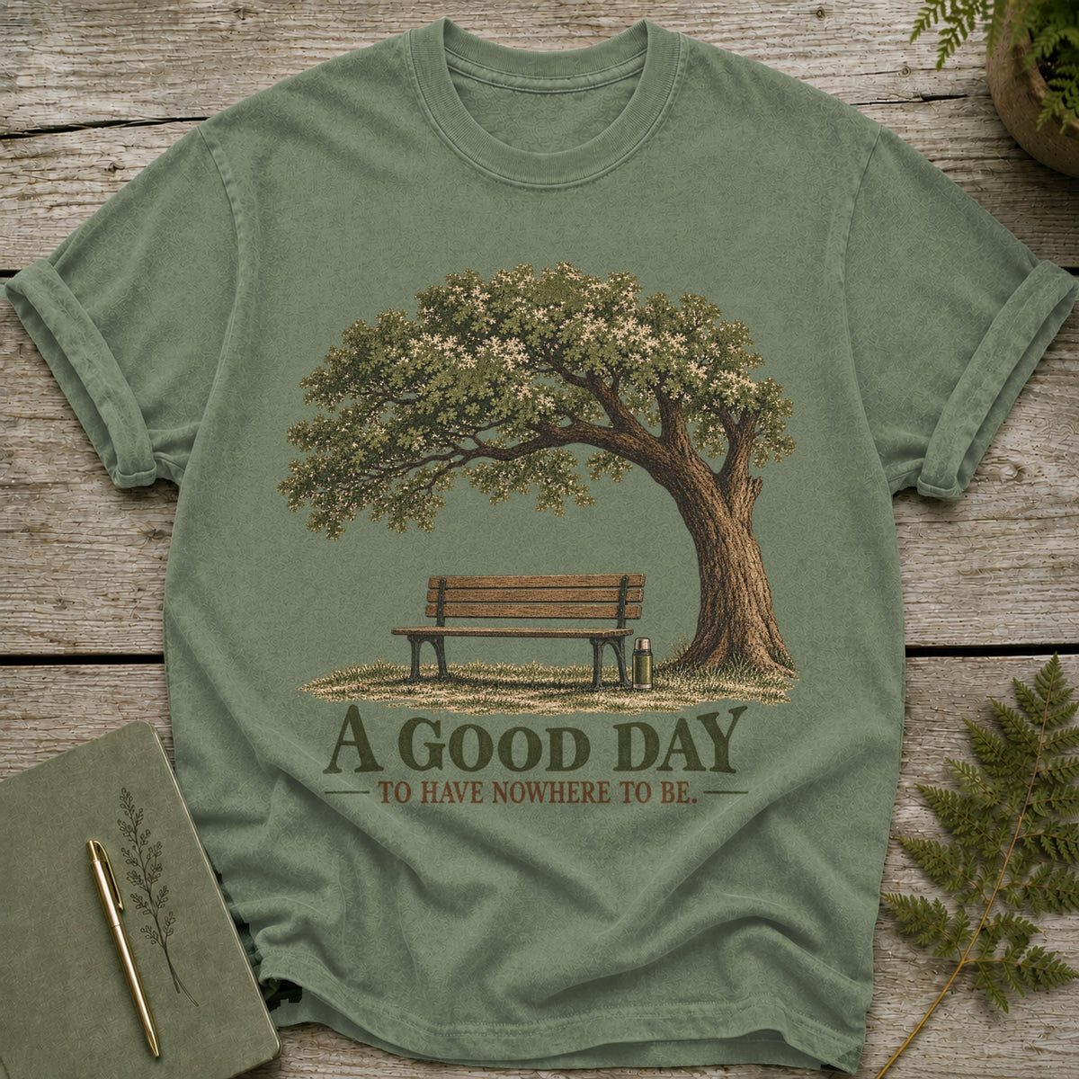 Green t-shirt with a graphic of a tree, bench, and thermos, featuring text.