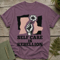 Purple t-shirt with graphic of hand holding flower and text "SELF CARE IS REBELLION".