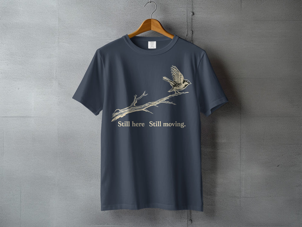 Dark grey t-shirt with a bird on a branch and text: "Still here Still moving."