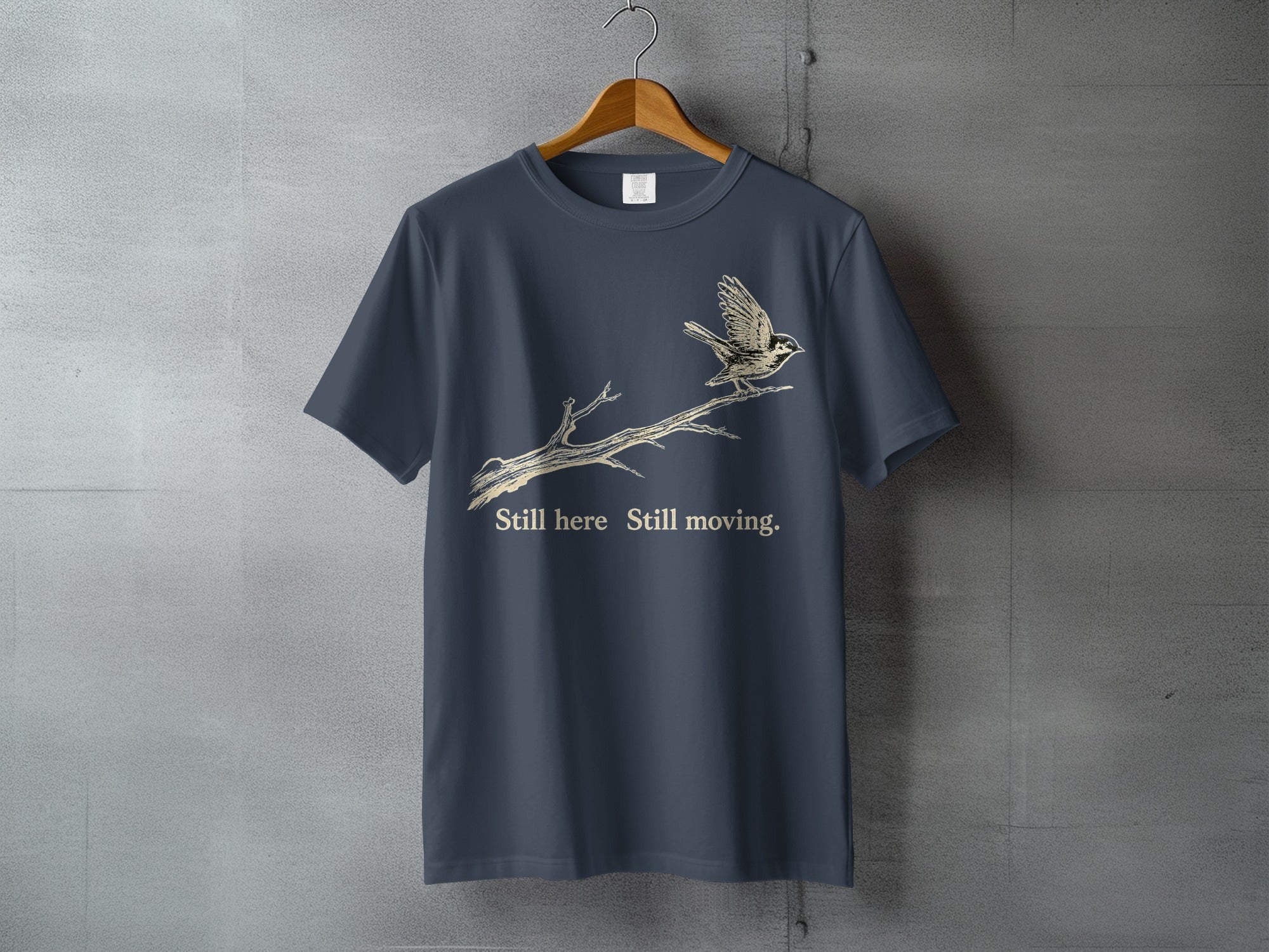 Dark grey t-shirt with a bird on a branch and text: "Still here Still moving."