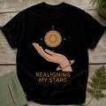 Black t-shirt with a hand holding a sun graphic and text "REALIGNING MY STARS".