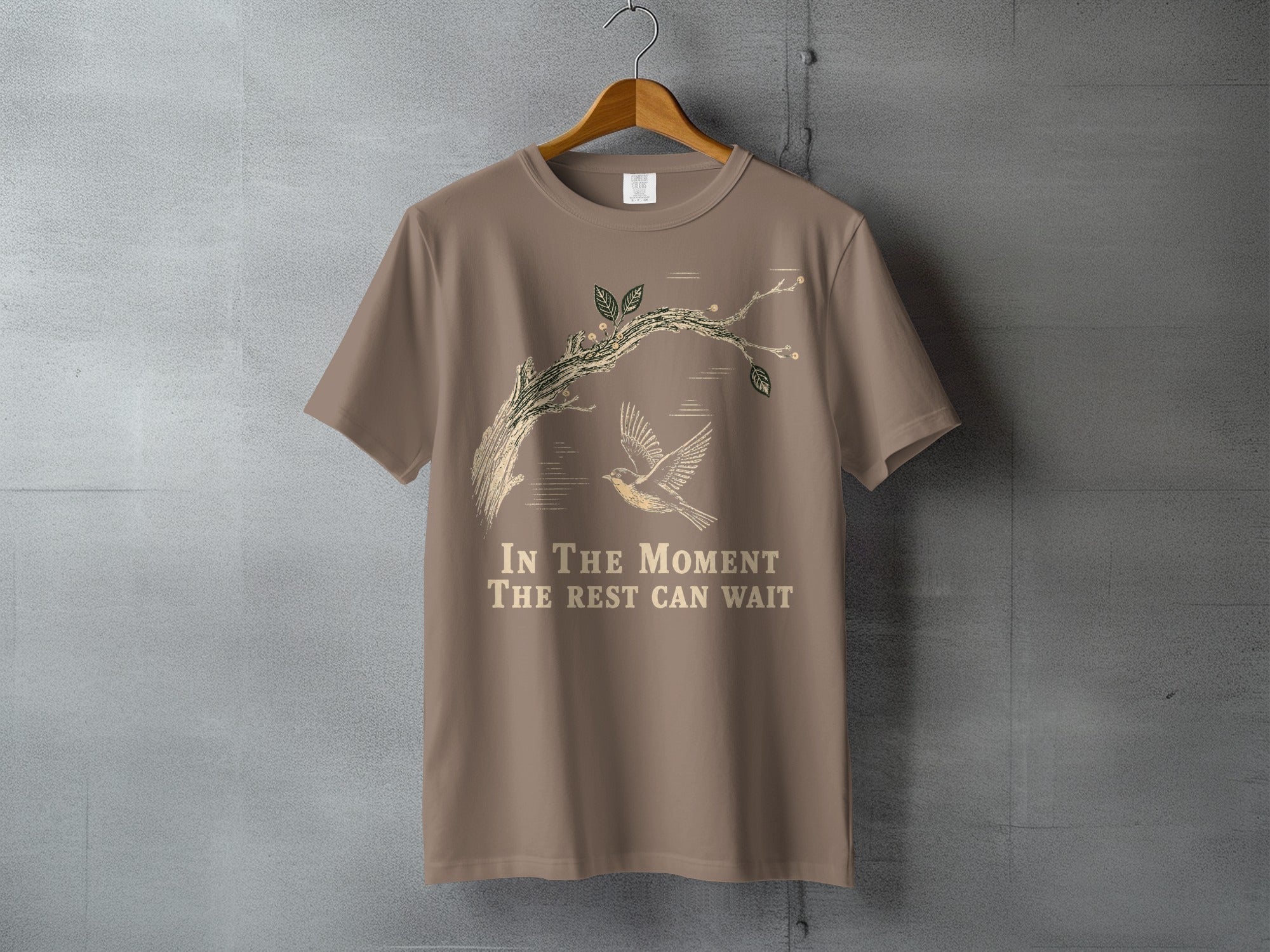 Taupe t-shirt with a bird and branch graphic and text "In The Moment The Rest Can Wait".