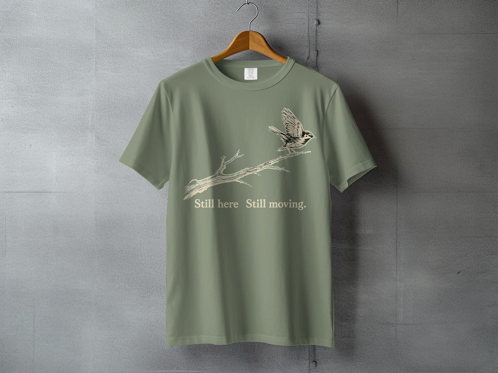 Olive green t-shirt with bird on branch graphic and text "Still here Still moving."