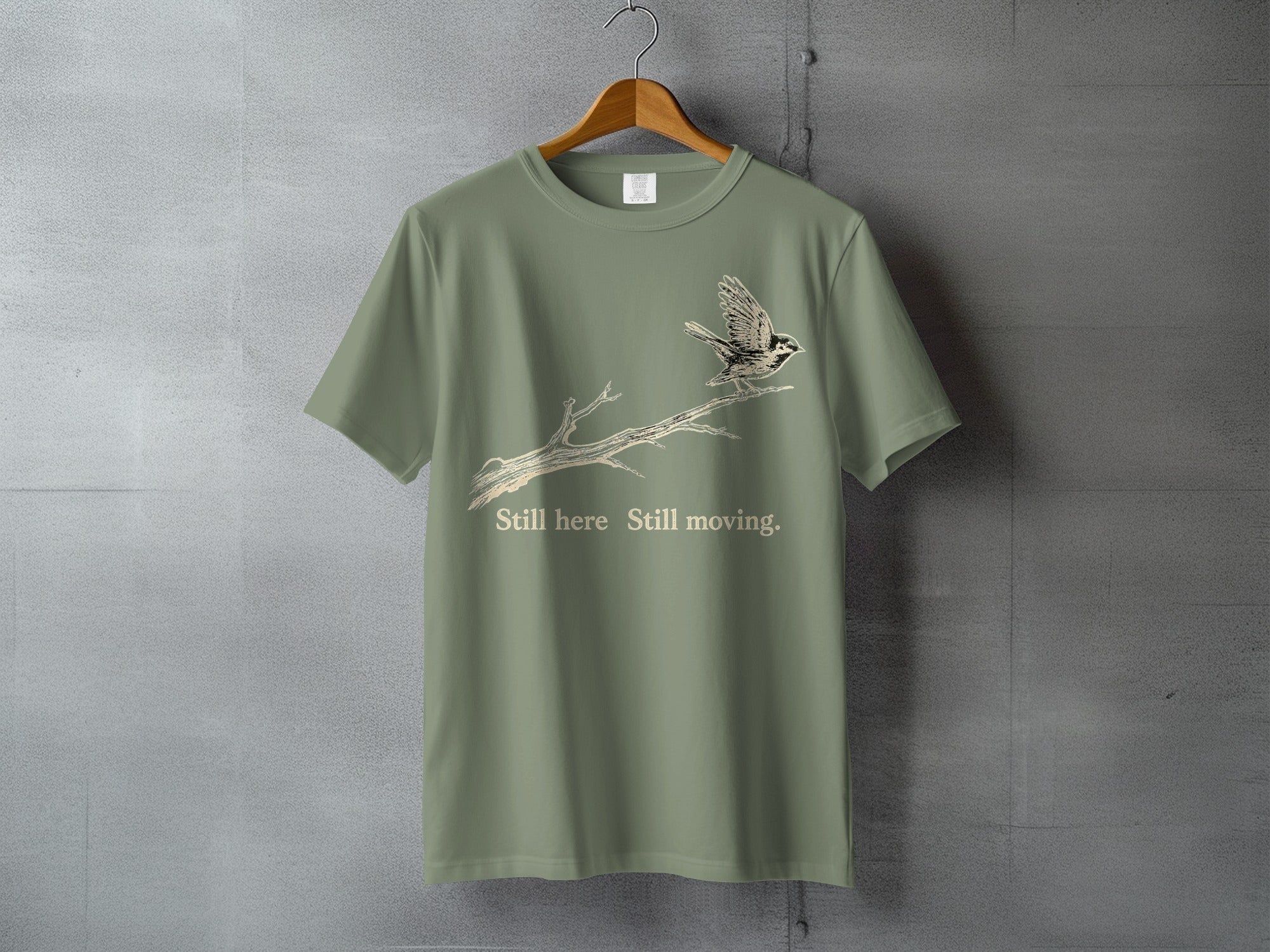 Olive green t-shirt with bird on branch graphic and text "Still here Still moving."
