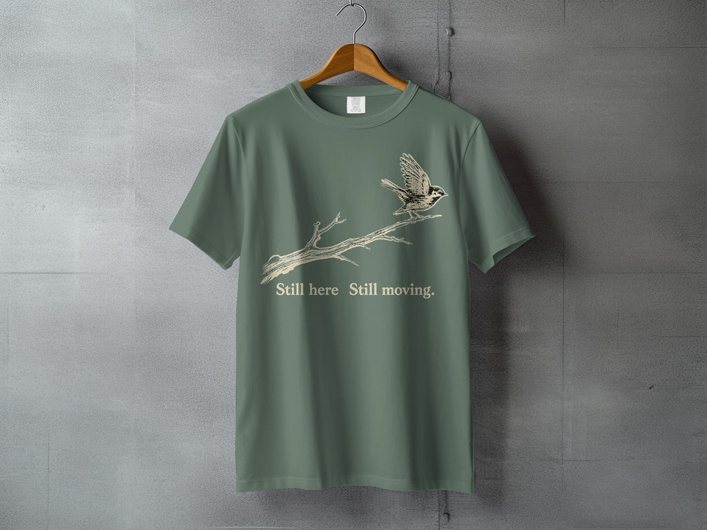Green t-shirt with a bird on a branch graphic and text "Still here Still moving."