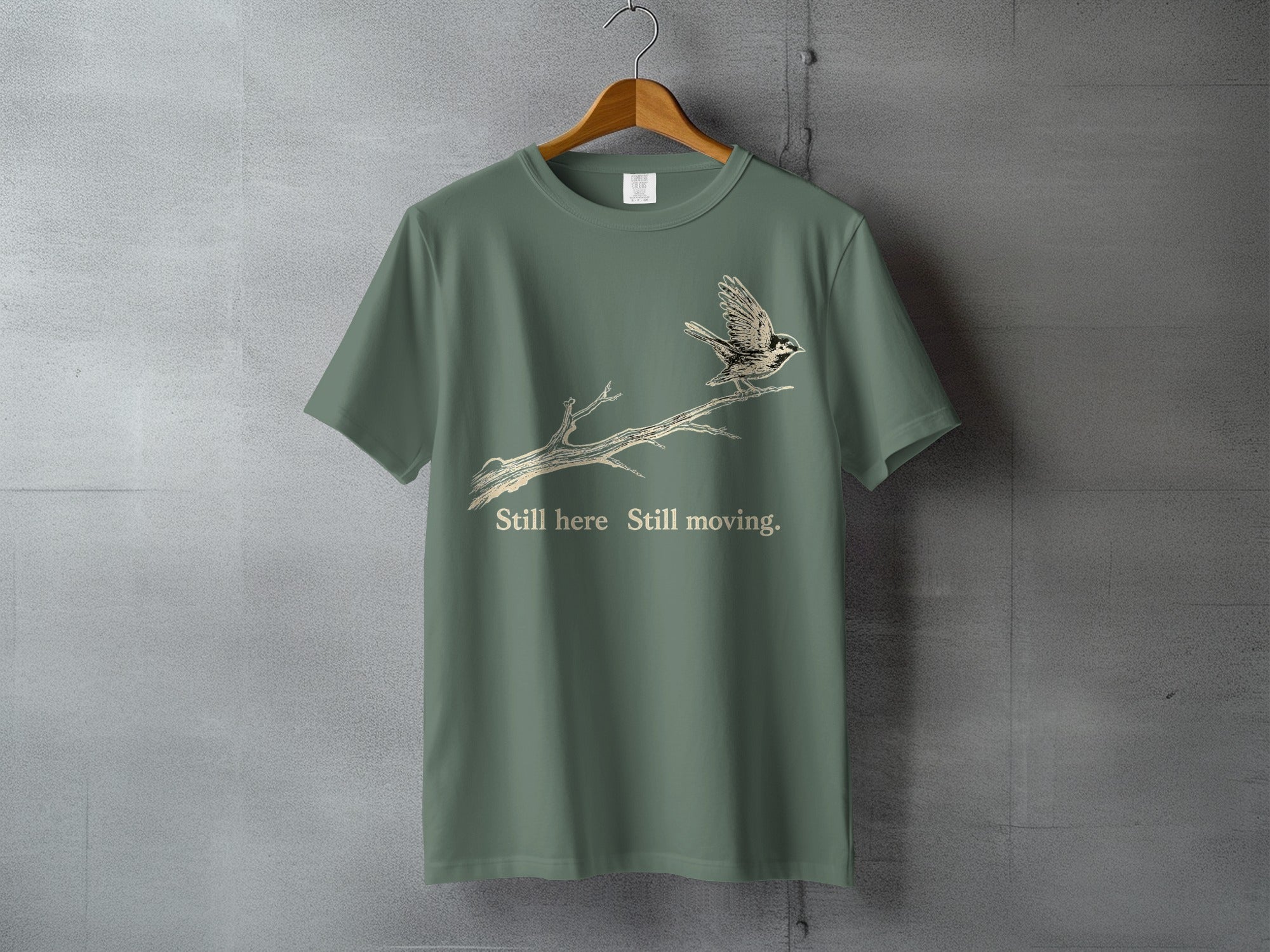 Green t-shirt with a bird on a branch graphic and text "Still here Still moving."
