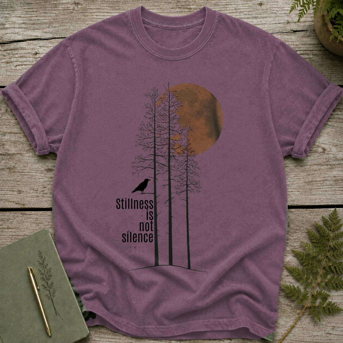 Purple t-shirt with crow, trees, and moon graphic with text "Stillness is not silence."