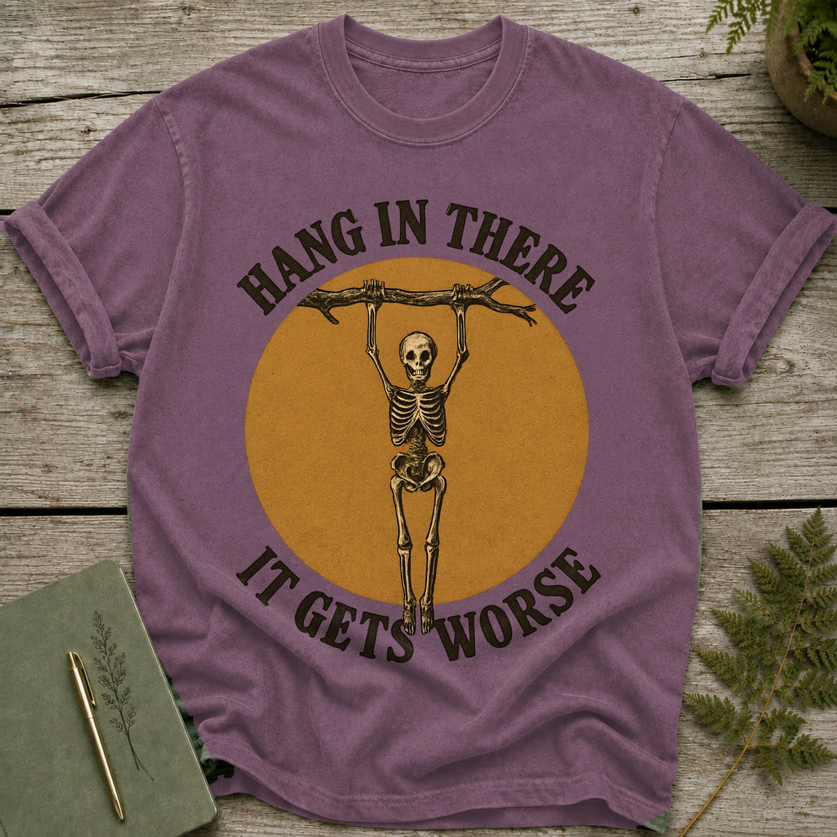 Purple t-shirt with skeleton hanging from branch, text "Hang in there it gets worse."