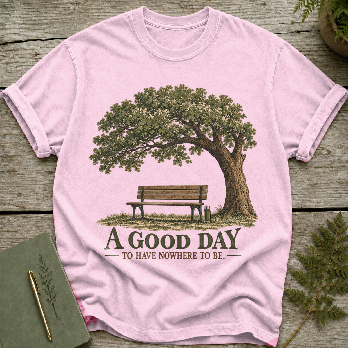 Pink t-shirt with a graphic of a tree, bench, and thermos, and text "A GOOD DAY TO HAVE NOWHERE TO BE."