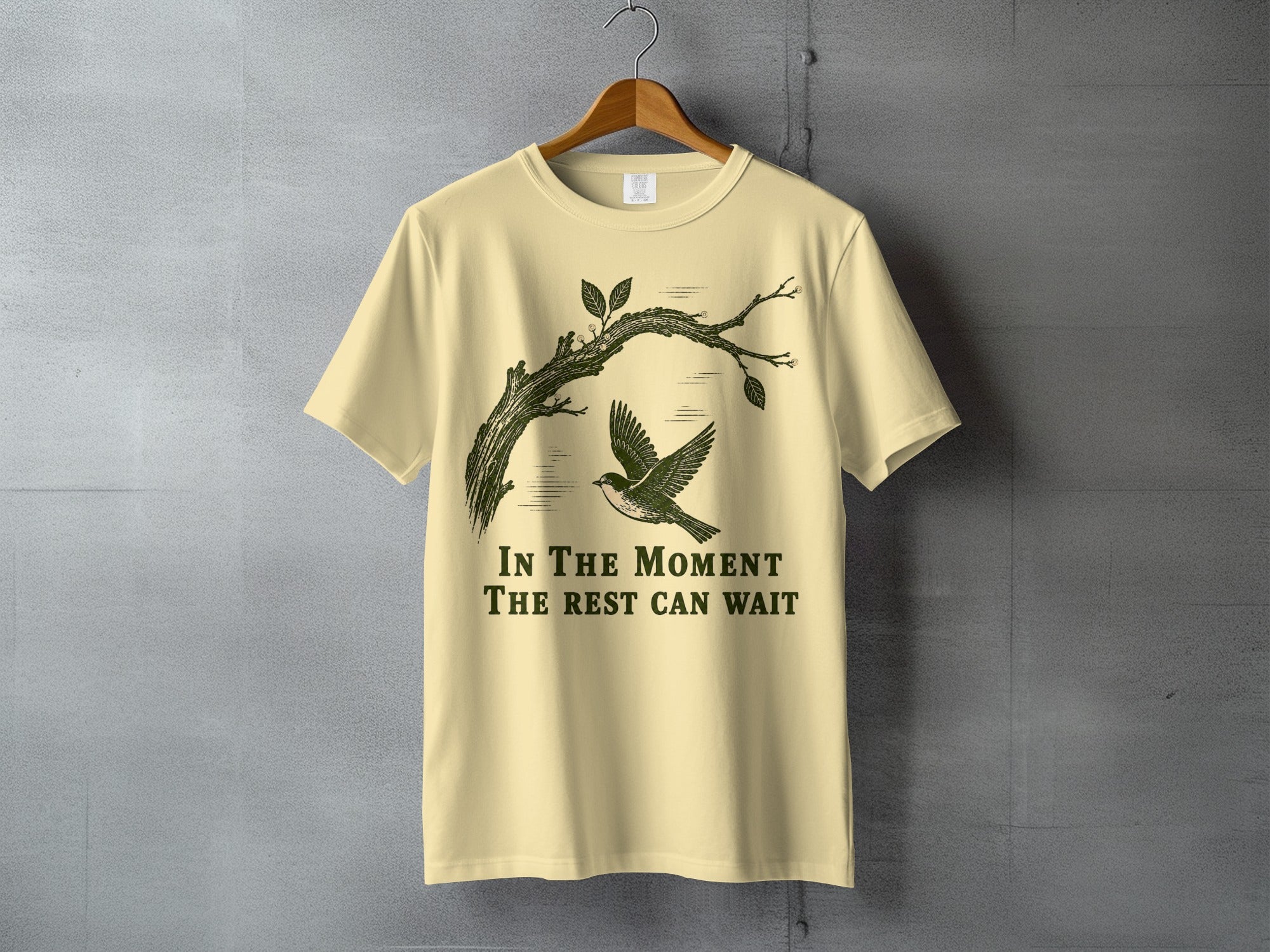 Light yellow t-shirt with a bird on a branch and the text "In The Moment The Rest Can Wait".