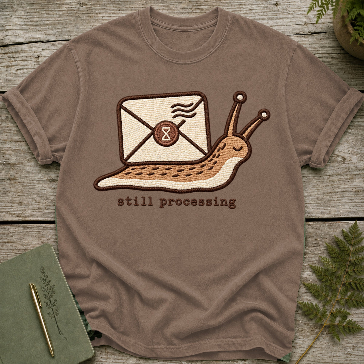 Brown t-shirt with embroidered slug carrying an envelope, saying "still processing".