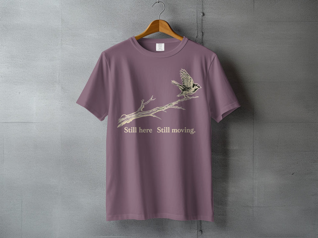 Mauve t-shirt with a bird on a branch graphic and text "Still here Still moving."