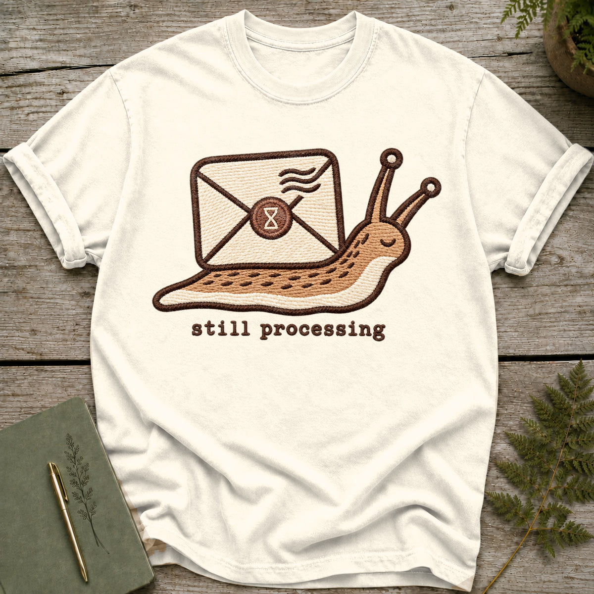 Cream t-shirt with embroidered snail carrying an envelope and the text "still processing".