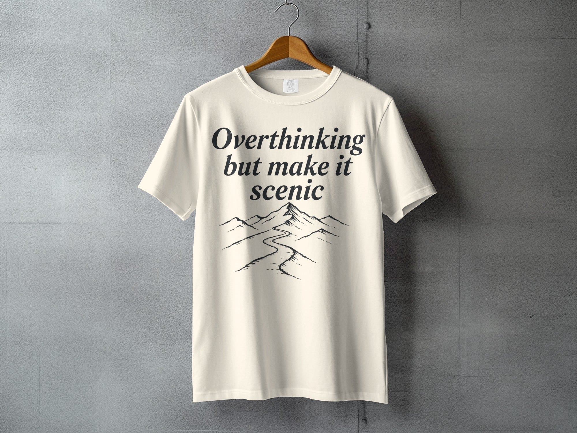 Cream t-shirt with "Overthinking but make it scenic" text and mountain landscape graphic.