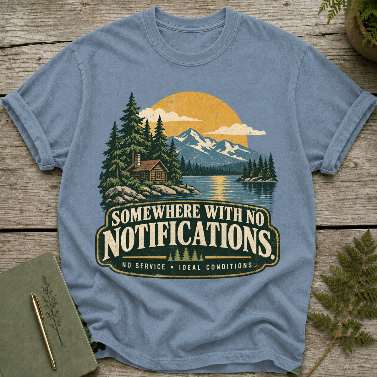 Blue t-shirt graphic with mountains, lake, cabin, trees, and text.