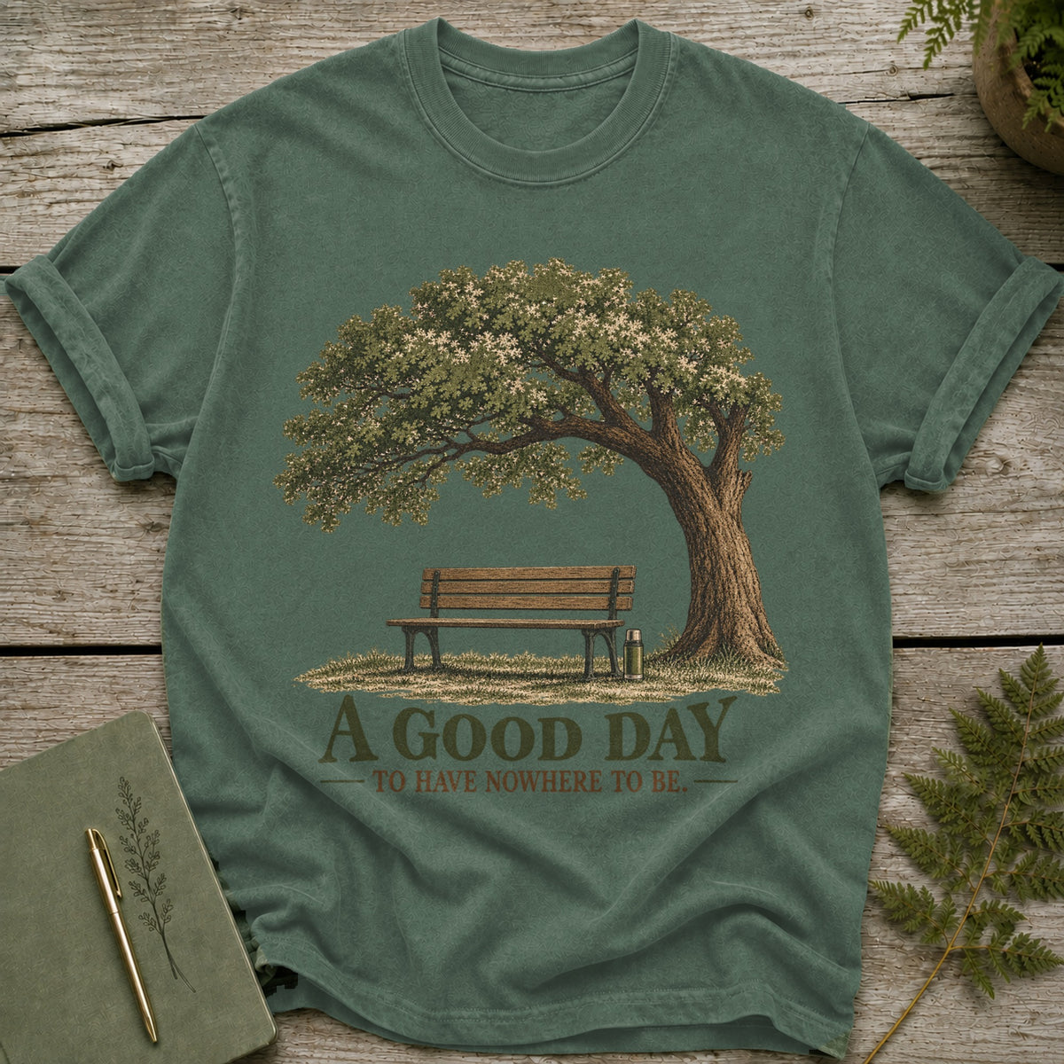 Green t-shirt with graphic of a tree, bench, and text "A GOOD DAY TO HAVE NOWHERE TO BE."