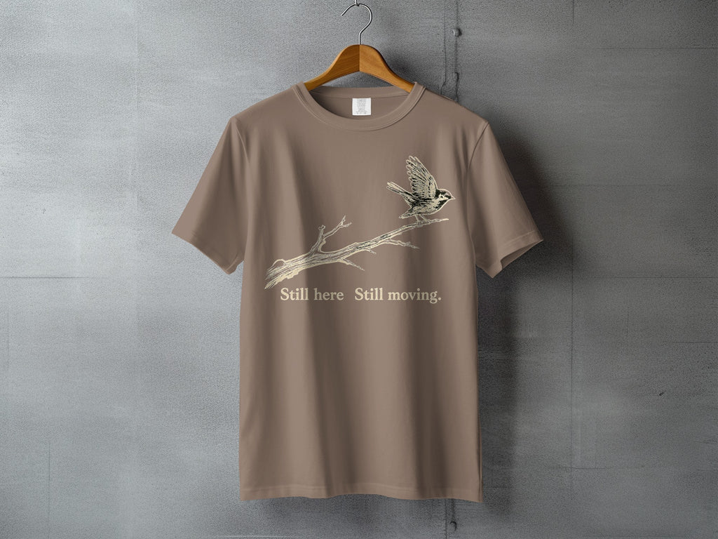 Taupe t-shirt with bird on branch graphic and text "Still here Still moving."