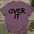 Purple t-shirt with black "OVER IT" text, laid flat on a wooden surface with greenery.